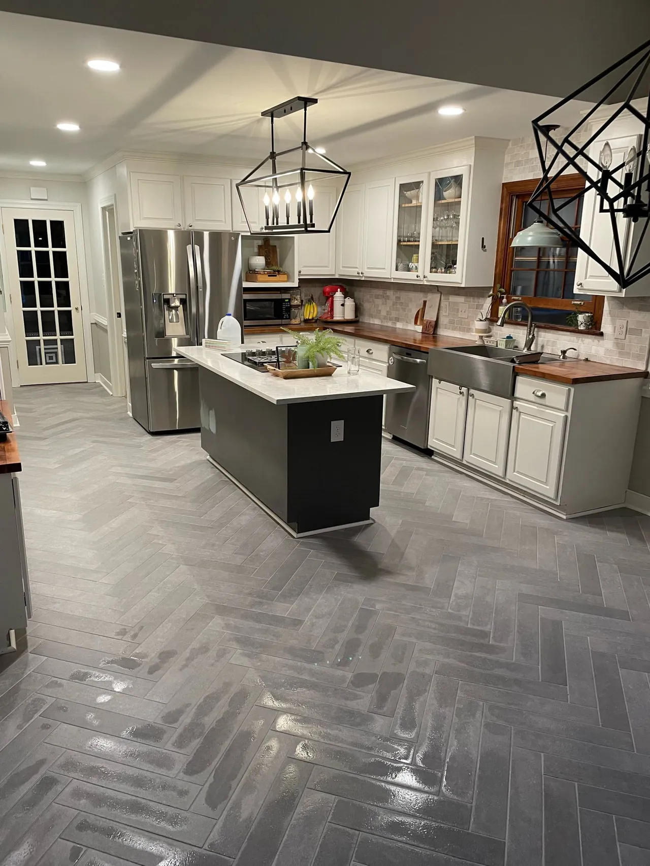 Wide view of a kitchen with a gray herringbone tile floor, white cabinets, a dark island with a white top and a farmhouse sink